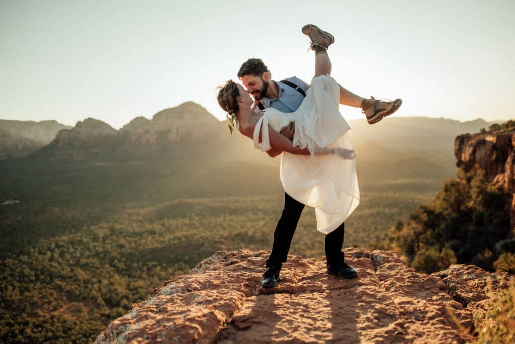 groom holding bride on edge of cliff, both smiling. Brides legs are up in the air showing her hiking boots under her dress.