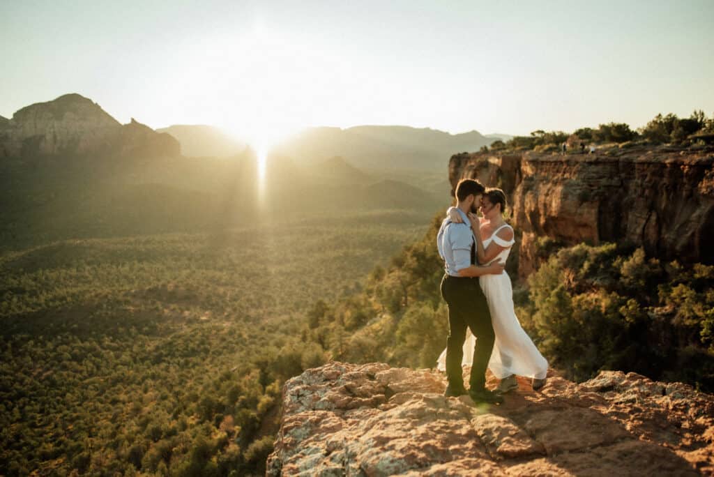 Bride and Groom holding each other with foreheads together on cliff edge with sunrise behind.