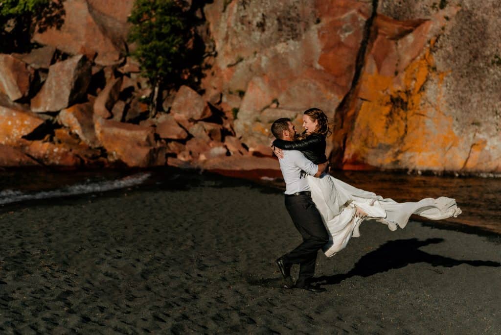 Groom swinging his bride around in front of red cliffs on Minnesota's black beach