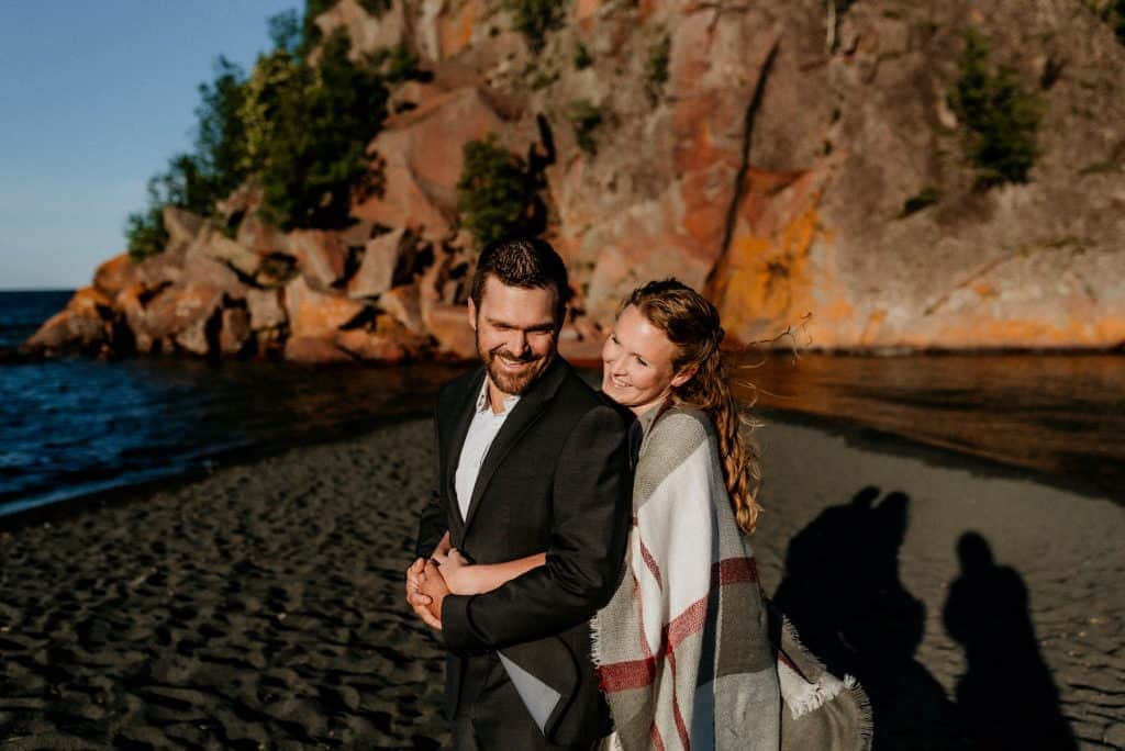 bride hugging groom on black sand each with red cliffs and Lake Superior behind 