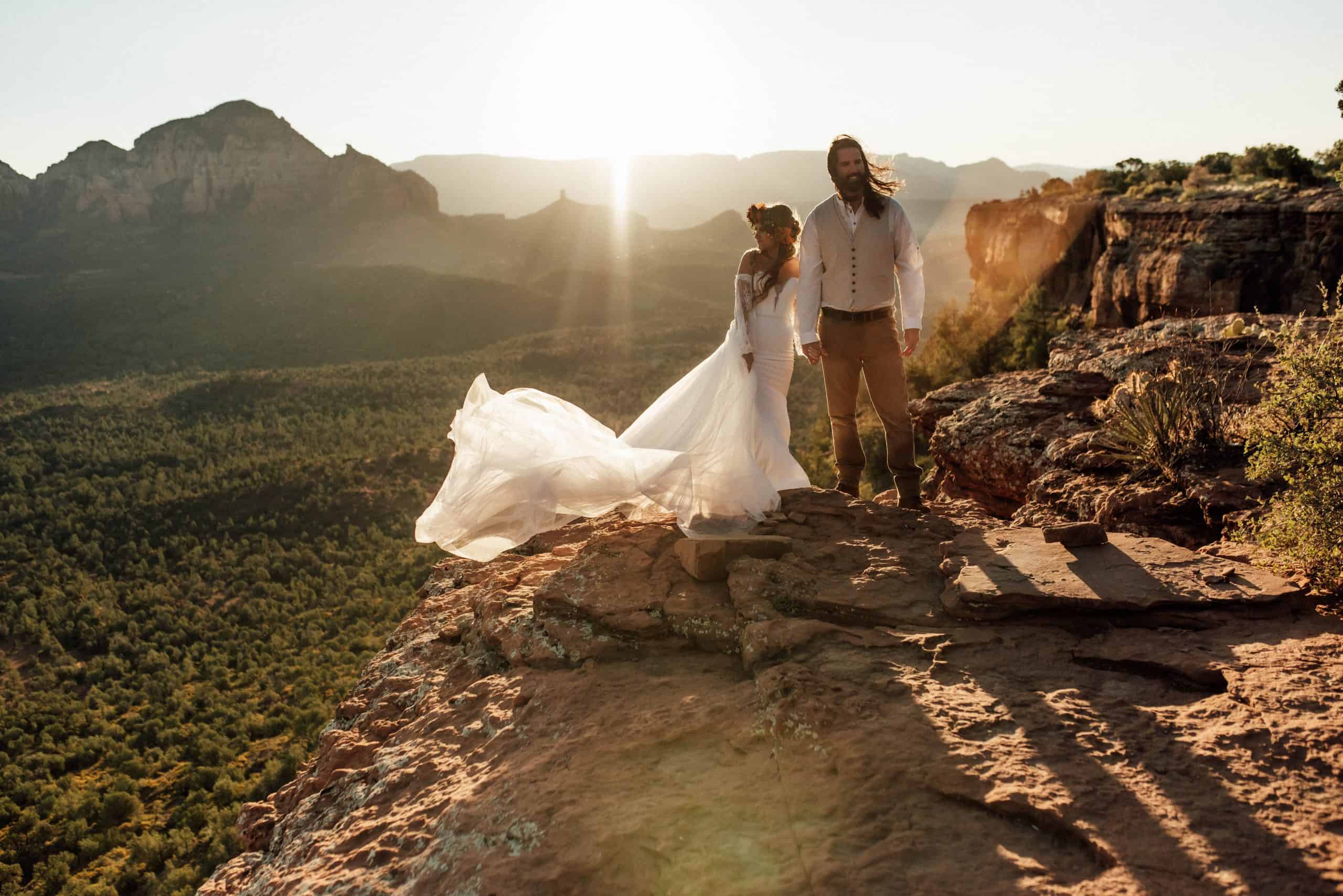 Elopement couple in neutral colors on cliff edge in Sedona, AZ with sunrise behind. Brides dress is blowing in the wind.